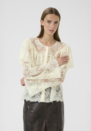 Blusa in pizzo a maniche lunghe in un morbido avorio con una texture traslucida, maniche arricciate e orlo scallop. Abbinata a una gonna in pelle marrone.
