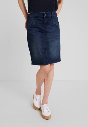 Personne portant une jupe en jean bleu foncé longueur genou avec poches avant, un haut noir et des baskets blanches à plateforme, debout les jambes croisées.