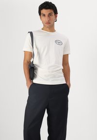 White cotton t-shirt with black graphic logo on the left side, paired with dark trousers. Model holds a small black bag.