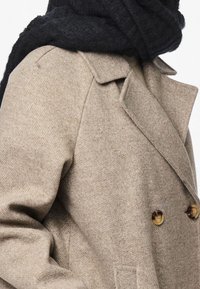 Manteau beige croisé en tissu texturé, doté de grands boutons dorés, de revers crantés et d'une écharpe noire drapée autour du cou.