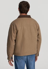 Mężczyzna w brązowej kurtce z ciemnym kołnierzem oraz niebieskich dżinsach, stojący tyłem na jednolitym jasnym tle.