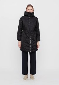 Cappotto puffo nero con finitura lucida, colletto alto e chiusura con cerniera. Presenta maniche lunghe e lunghezza fino a metà coscia. Indossato sopra pantaloni scuri.