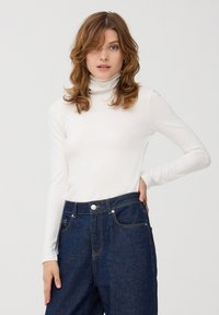 Biała bluzka z długim rękawem i golfem, wykonana z gładkiego materiału, zestawiona z ciemnoniebieskimi dżinsami z wysokim stanem, wyposażonymi w kieszenie i złotymi przeszyciami.