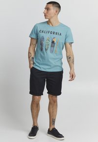 Plava pamučna majica s natpisom "CALIFORNIA" i grafikom daske za surfanje. Uparena s tamnim kratkim hlačama i crnim tenisicama.