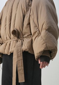 Veste matelassée marron surdimensionnée avec ceinture nouée à la taille, portée sur une jupe noire, avec une main visible sur le côté.