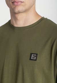 Camiseta de manga corta en verde oliva hecha de algodón, con un escote redondo y un parche de logo negro texturizado en el lado izquierdo del pecho.