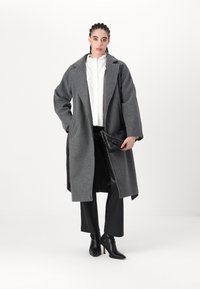 Grau Wolle Übermantel mit lockerem Schnitt, großen Revers, kombiniert mit einer weißen Rüschbluse und schwarzen maßgeschneiderten Hosen. Schwarze Handtasche und Ankle Boots.