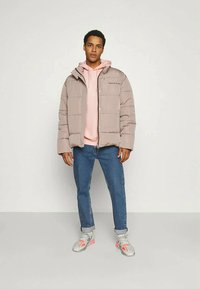 Chaqueta acolchada marrón con capucha, cierre de botones, llevada sobre una sudadera rosa clara. Combinada con jeans azules y zapatillas grises con acentos rosas.