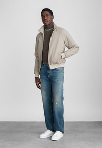 Μπεζ ζακέτα με φερμουάρ, φτιαγμένη με ραβδωτό γιακά, συνδυασμένη με καφέ πλεκτό πουλόβερ και ανοιχτό μπλε τζιν με φθορές. Οι λευκές αθλητικές μπότες ολοκληρώνουν το ντύσιμο.