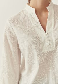 Chemise en lin blanc avec motifs de feuilles brodés, dotée d'une patte de boutonnage avec de petits boutons et d'un col mao. Texture douce et légère.