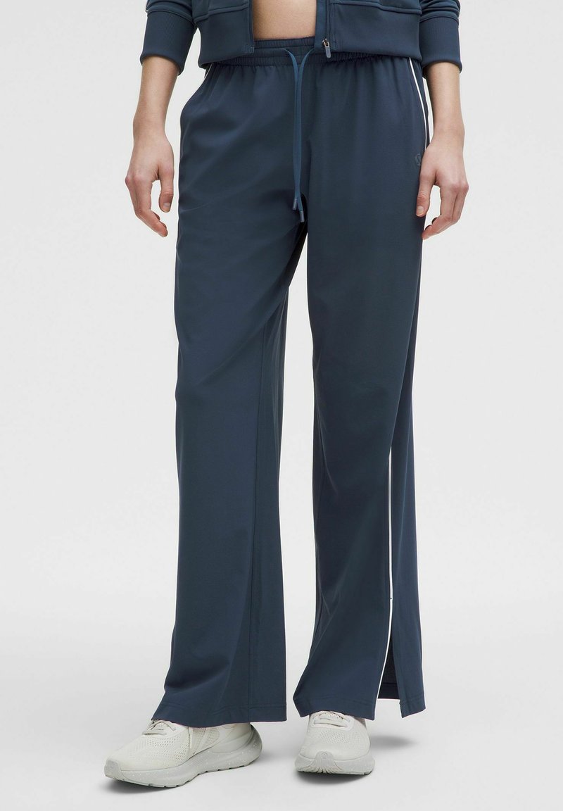 Mornarske sportske hlače s elastičnom doradom u struku, vezicom i bočnim prorezima. Izrađene od glatkog, laganog materijala. Uparene s bijelim tenisicama.