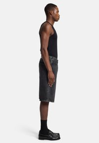Canotta nera, pantaloni shorts in denim grigio scuro, vista laterale. Le caratteristiche includono due tasche posteriori, passanti per cintura e vestibilità rilassata. Calzature: mocassini neri.