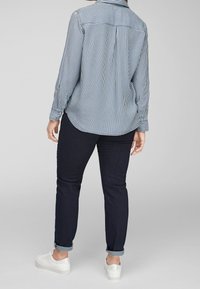 Camicia a strisce blu e bianche, vista posteriore. Abbinata a jeans di denim scuro, con risvolti e sneakers bianche. Design semplice e moderno.