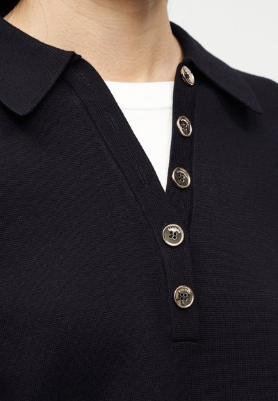 Κοντινό πλάνο ενός μαύρου πουκαμίσου με γιακά και πέντε χρυσά κουμπιά πάνω από ένα λευκό εσώρουχο, στο πάνω μέρος του στήθους και του λαιμού ενός ατόμου.