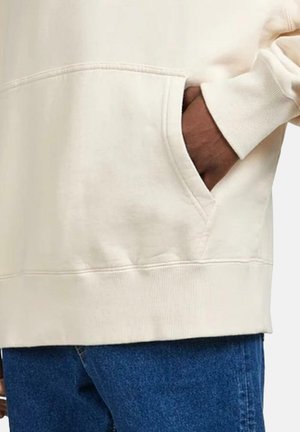 Persoon draagt een crèmekleurige trui met de hand in de voorzak en een blauwe spijkerbroek zichtbaar onder de zoom van de trui.