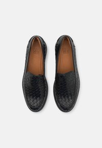 Černé pletené kožené slip-on boty s hladkým hnědým interiérem a kulatou špičkou, s texturovanými akcenty a plochou podrážkou.