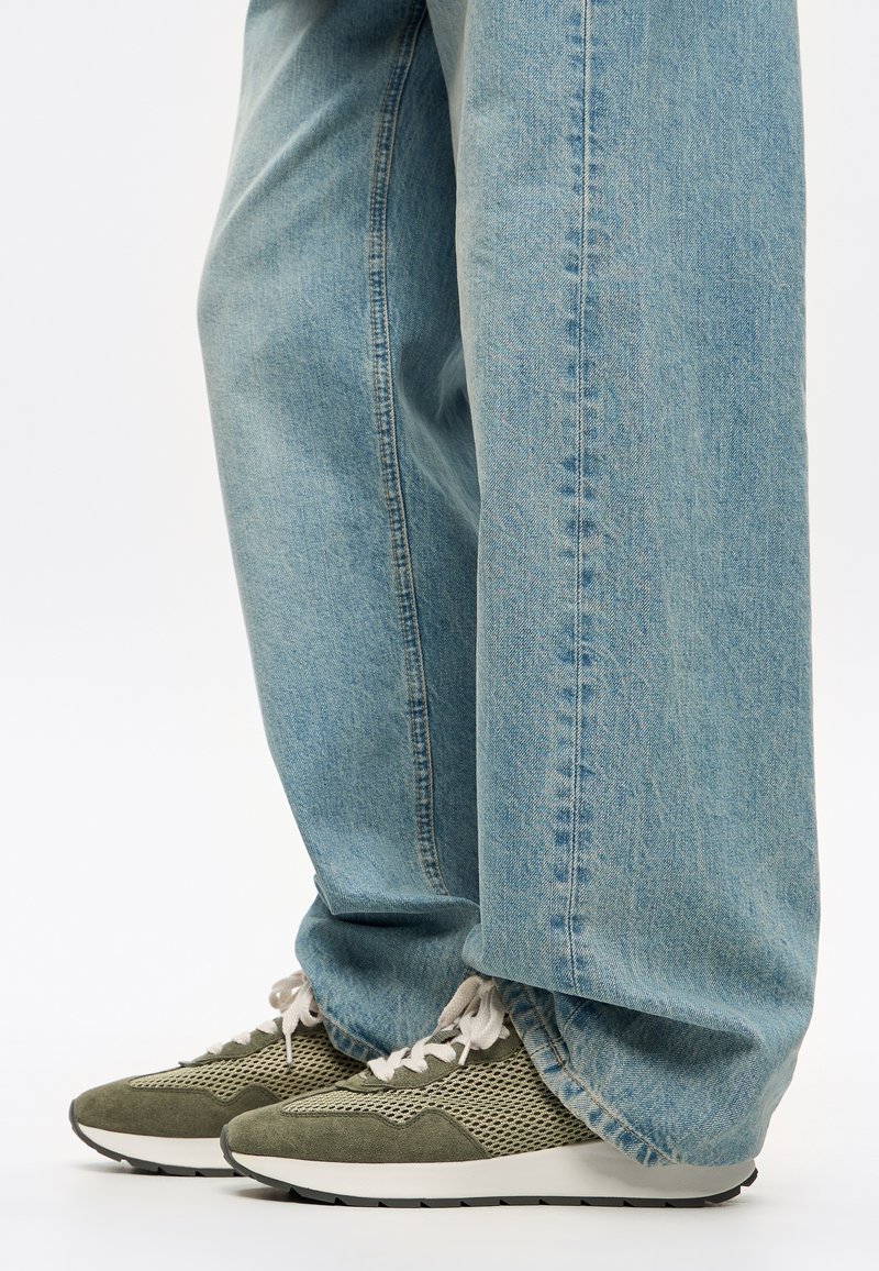 Jeans azzurri chiari con vestibilità ampia, abbinati a sneakers in mesh verde e suede con suola bianca e lacci grigio chiaro.