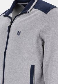 Lichtgrijze zip-up hoodie met marineblauwe accenten op de schouders, het ritsvak, de binnenzijde van de capuchon en een klein gevleugeld logo op de borst.