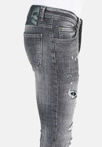 Slanke grijze denim jeans met verweren details, zichtbare scheuren en een gestructureerd oppervlak. Heeft een zwarte leren patch op de tailleband.