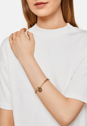 Gul kædearmbånd med en rund charm, der har et struktureret design. Bæret på en bar arm, mod en hvid t-shirt baggrund.