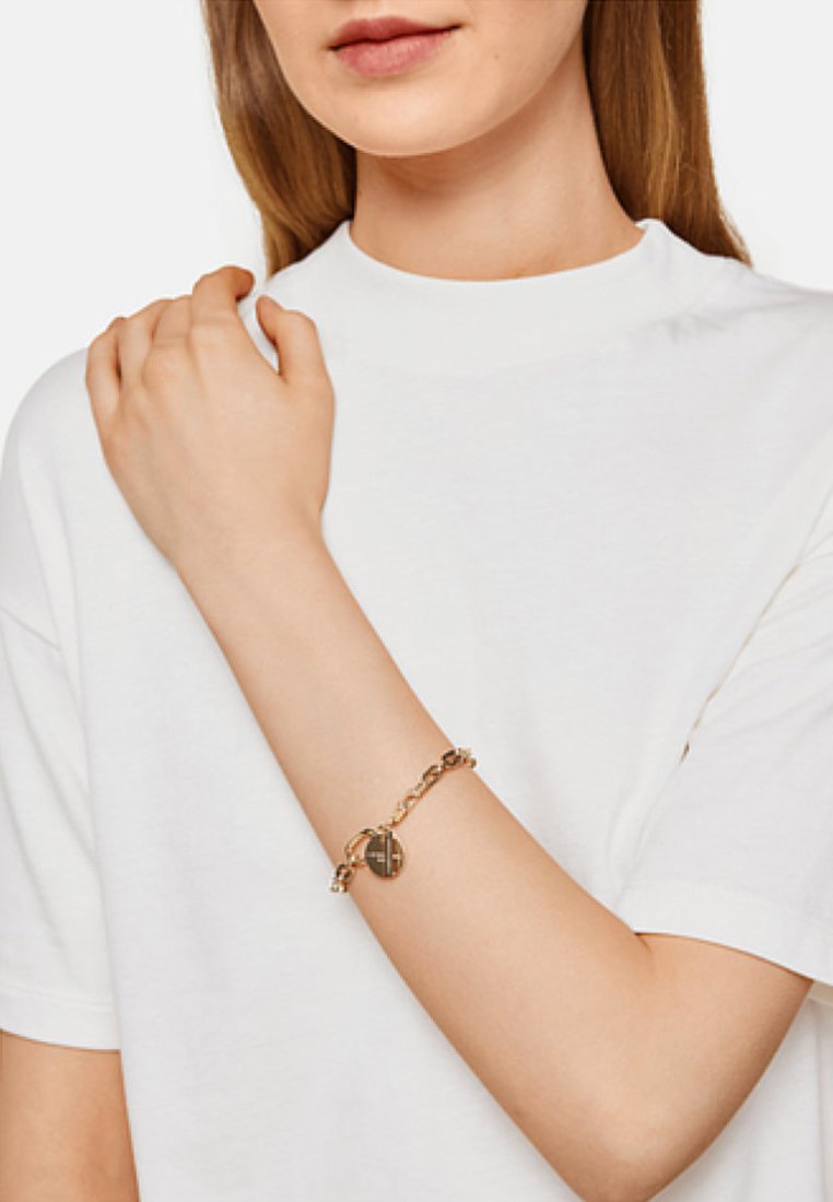 Guldkedjearmband med en rund berlock, som har en texturerad design. Buret på en bar arm, mot en vit t-shirt bakgrund.