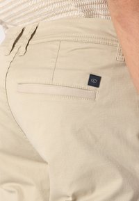 Beige Hose aus strukturiertem Stoff. Verfügt über eine einzelne Gesäßtasche mit einem kleinen blauen Logo-Tag und Gürtelschlaufen entlang der Taille.