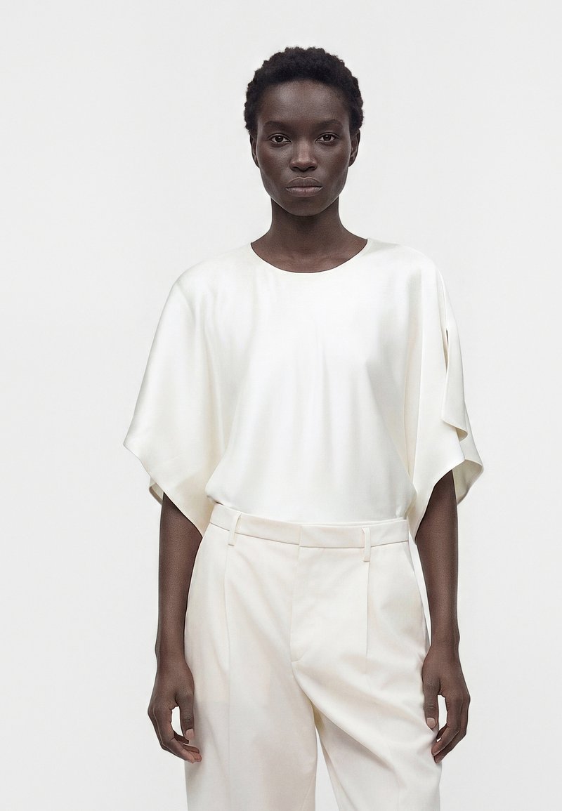 Femme aux cheveux courts et bouclés portant un chemisier blanc en satin oversize et un pantalon blanc taille haute, debout devant un fond uni.