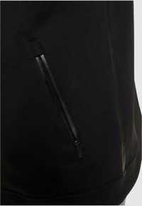 Sort sweatshirt med en glat tekstur, sidelomme med lynlås i sort og en afslappet pasform. Detaljerne inkluderer minimal syning.