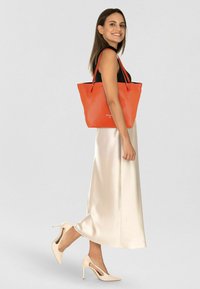 Oranje leren handtas met een gestructureerd oppervlak, zwart interieur en twee schouderriemen, weergegeven naast een lichtkleurige satijnen rok.