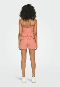 Lyserød ruffle romper med tynde stropper, der har en tekstureret stof og kort længde. Kombineret med hvide sneakers, set fra bagsiden.