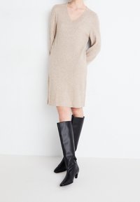 Beige stickad klänning med långa ärmar och V-ringning, kombinerad med knähöga svarta läderstövlar med spetsig tå och låg blockklack.