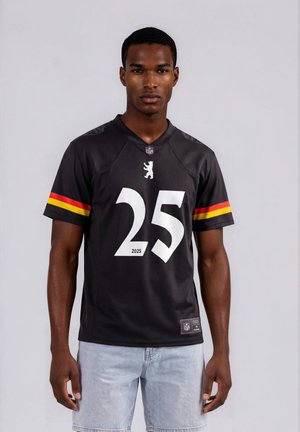 Jonge man draagt een zwart sportshirt met nummer 25 en geel-rode strepen op de mouwen, gecombineerd met lichtblauwe denimshorts, staand frontaal.