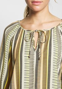Blouse multicolore avec des rayures verticales noires, jaunes et beiges. Elle a une encolure ronde avec un accent de noeud au centre.