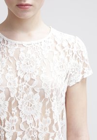 Detailní záběr osoby v bílém květinovém krajkovém topu s krátkým rukávem a kulatým výstřihem na jednobarvém světlém pozadí.