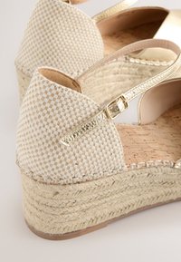 Närbild på beige vävda kilklacksandaler med flätad juteplattform och metalliskt guldfärgat ankelband med spänne.