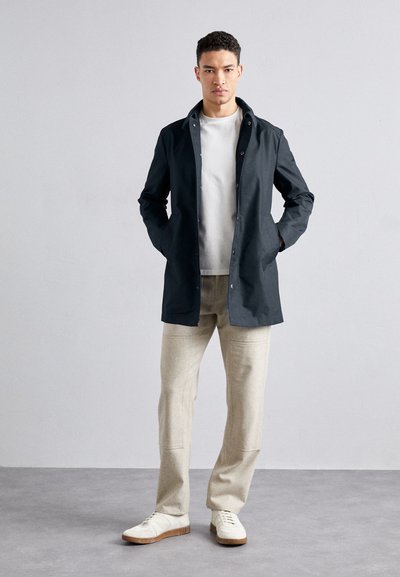 Dunkelblaue wasserdichte Jacke mit einem vorderen Druckknopfverschluss, Seitentaschen, getragen über einem hellgrauen T-Shirt und cremefarbenen Hosen. Weiße Sneaker.