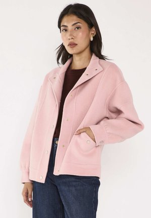 Femme aux cheveux foncés portant des boucles d'oreilles en perles, une veste rose boutonnée par-dessus un haut sombre et un jean bleu, posant avec une main dans la poche.