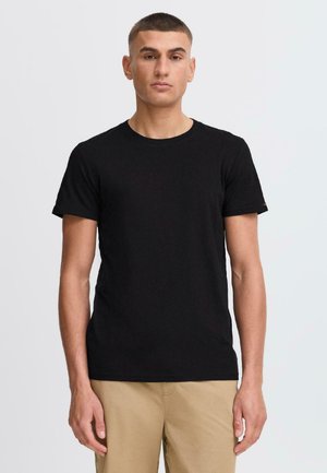 Camiseta de algodón negra con escote redondo y mangas cortas, de diseño ajustado, combinada con pantalones beige claros. Minimalista, sin patrones ni acentos visibles.