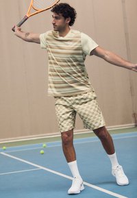 Muž v pruhovaném tričku a kostkovaných šortkách hraje uvnitř kurtu s oranžovou tenisovou raketou a kolem něj jsou rozházené tenisové míčky.