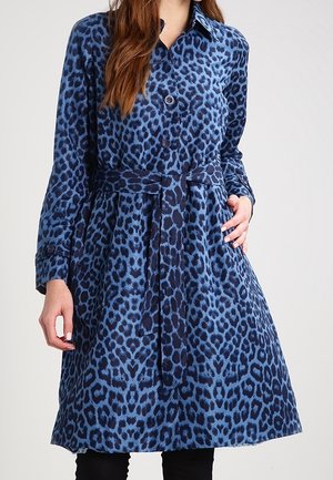 Kvinde iført en blå leopardprint kjole med bælte, lange ærmer og knappet foran, stående mod en ensfarvet lys baggrund.