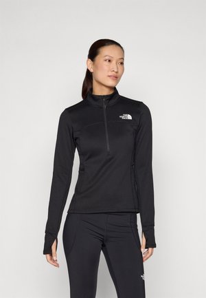 Kvinne iført svart langermet jakke med halv glidelås og leggings med The North Face-logoer, stående mot en ensfarget bakgrunn, ser mot høyre.