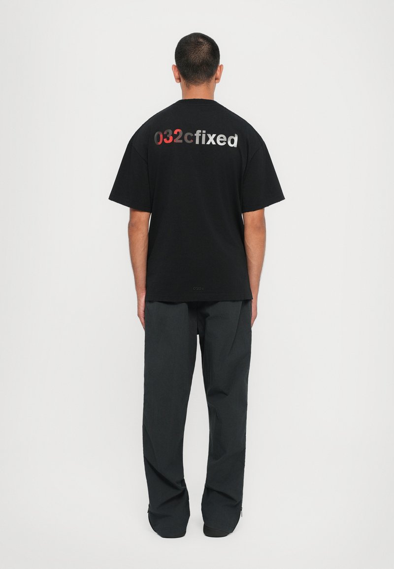 Schwarzes T-Shirt mit kurzen Ärmeln, auf dem Rücken mit "032cfixed" in Rot und Silber bedruckt; getragen mit dunkelgrauen Hosen.
