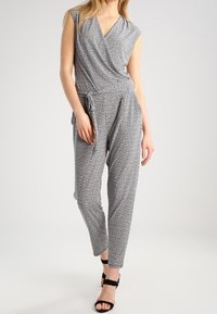 Combinaison à motifs noir et blanc avec des manches courtes, décolleté en wrap, taille cintrée et jambes taper à longueur de cheville. Équipée d'un détail à nouer.