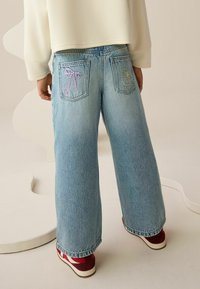 Jasnoniebieskie dżinsy o szerokich nogawkach z kolorowymi, haftowanymi detalami na tylnych kieszeniach i luźnym krojem. Połączone z czerwono-białymi trampkami.