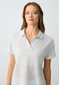 Jasnoniebieska koszulka polo z krótkim rękawem. Posiada kołnierzyk i pionowe paski. Wykonana z teksturowanej, przezroczystej tkaniny.