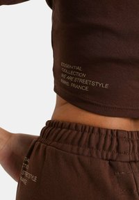 Brun cropped t-shirt og matchende sweatpants med elastisk talje. Gyldent trykt tekst: "ESSENTIAL COLLECTION VI ER STREETSTYLE PARIS, FRANKRIG."