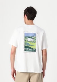 Witte katoenen t-shirt met een grafische afdruk die doet denken aan een postzegel met een illustratie van een golfclub, omrand met scalloped randen.