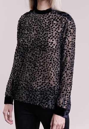 Nainen pitää yllään pitkähihaisen, läpikuultavan mustan topin, jossa on samettipilkkuja, aluskerroksena nude-värinen, yhdistettynä mustiin housuihin.