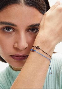 Due braccialetti a corda - uno nero e uno blu - presentano piccoli ciondoli geometrici. Il filo nero è intrecciato con quello blu, indossato su un polso.