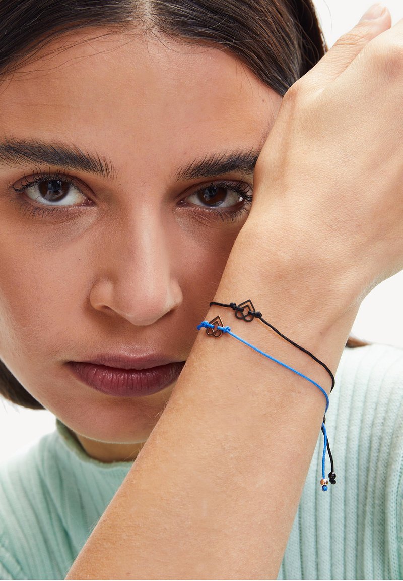 Due braccialetti a corda - uno nero e uno blu - presentano piccoli ciondoli geometrici. Il filo nero è intrecciato con quello blu, indossato su un polso.